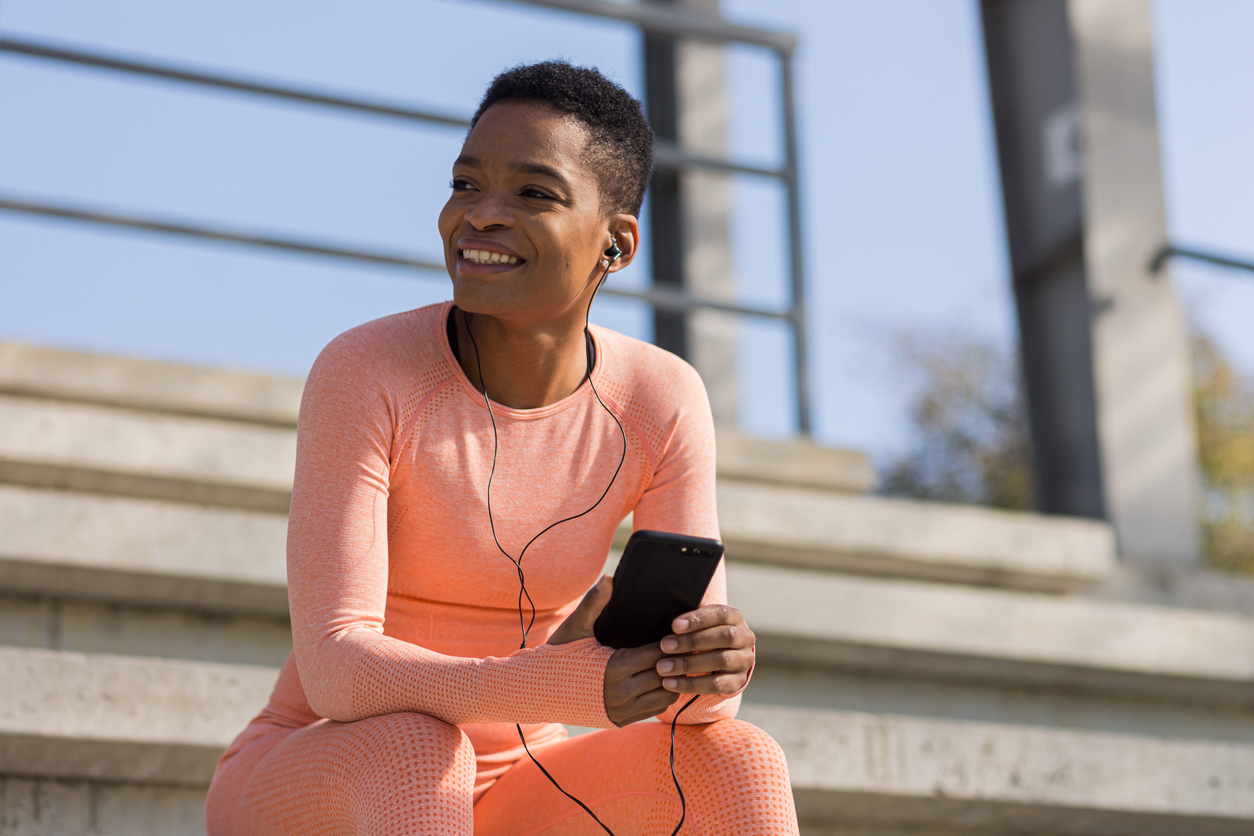 Beautiful and successful female athlete smiling listens to audio book from mobile phone during morning fitness and jogging near stadium, african american woman with mobile phone